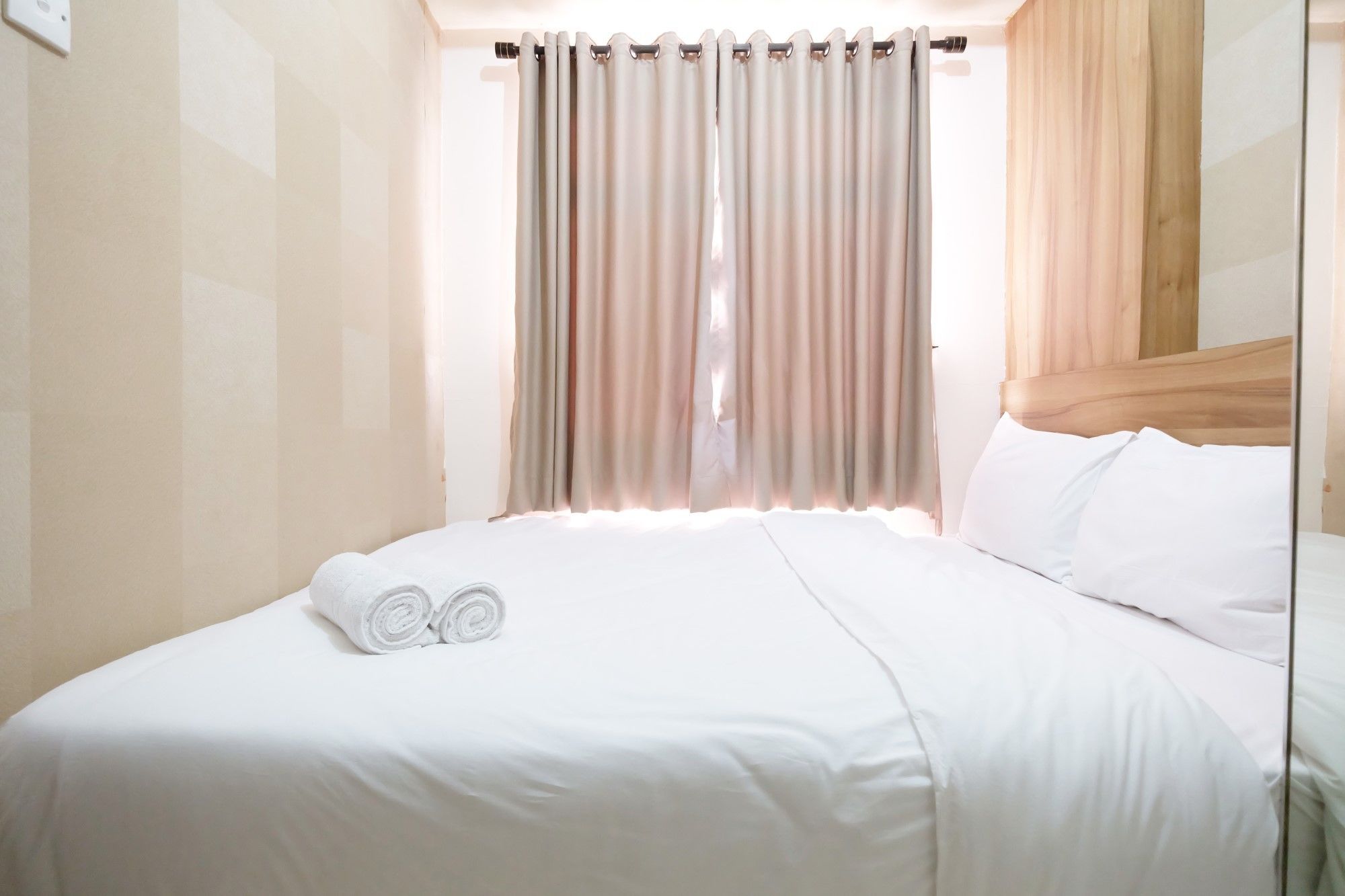 A neatly made bed with white linens and two rolled towels, a wooden headboard, and a window with beige curtains.