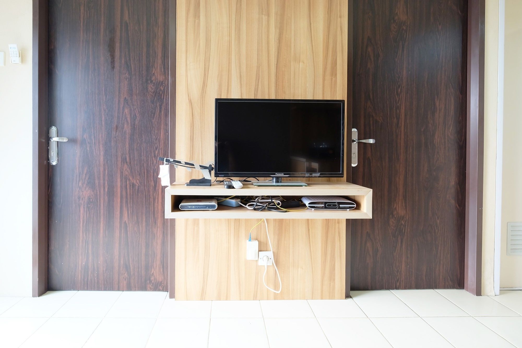 A flat-screen TV mounted on a wooden cabinet with a shelf below, positioned between two dark wooden doors.