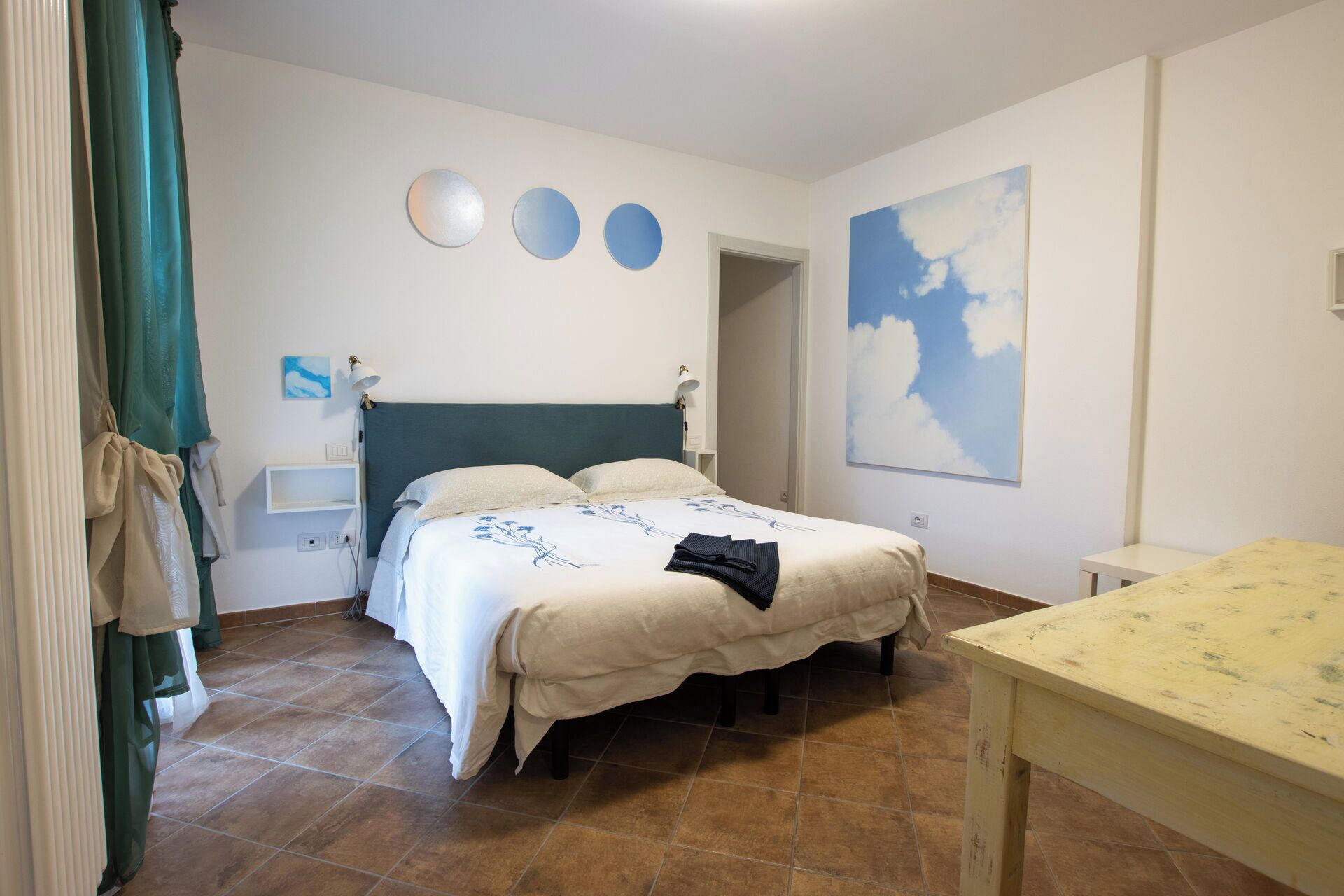 Une chambre à coucher avec un lit, des tables de chevet, un bureau et un tableau représentant des nuages accroché au mur.