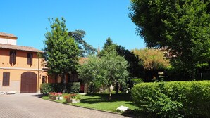 A courtyard with a paved walkway, a building with a tiled roof, and lush greenery. - Giardino 34 (Forlì)