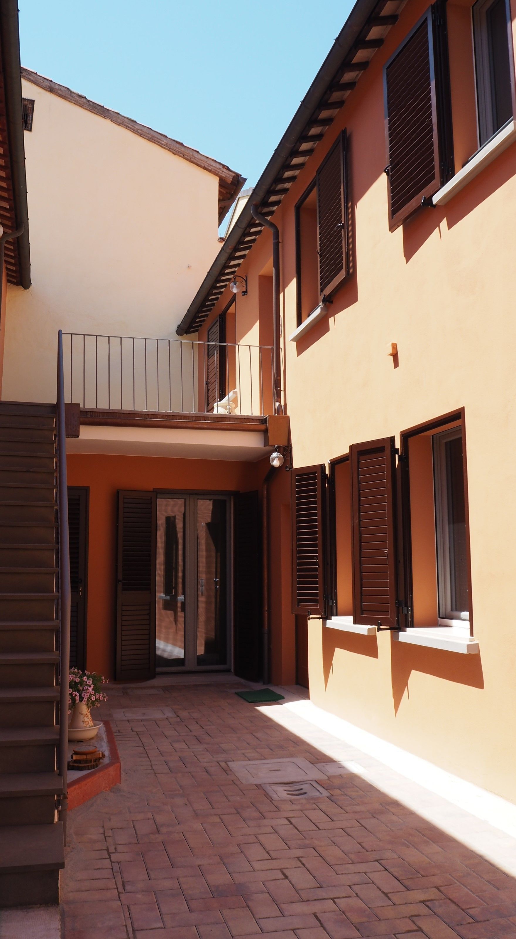 A courtyard with a brick floor, a small garden with flowers, and a building with multiple windows and shutters.