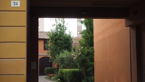 A view through an open doorway into a courtyard with a building featuring a clock tower, surrounded by greenery and a paved path. - Giardino 34 (Forlì)