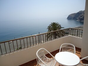 A balcony with white wicker chairs and a small round table, overlooking a beach with a clear view of the sea and a palm tree. - Hotel Las Conchas (Albunol)