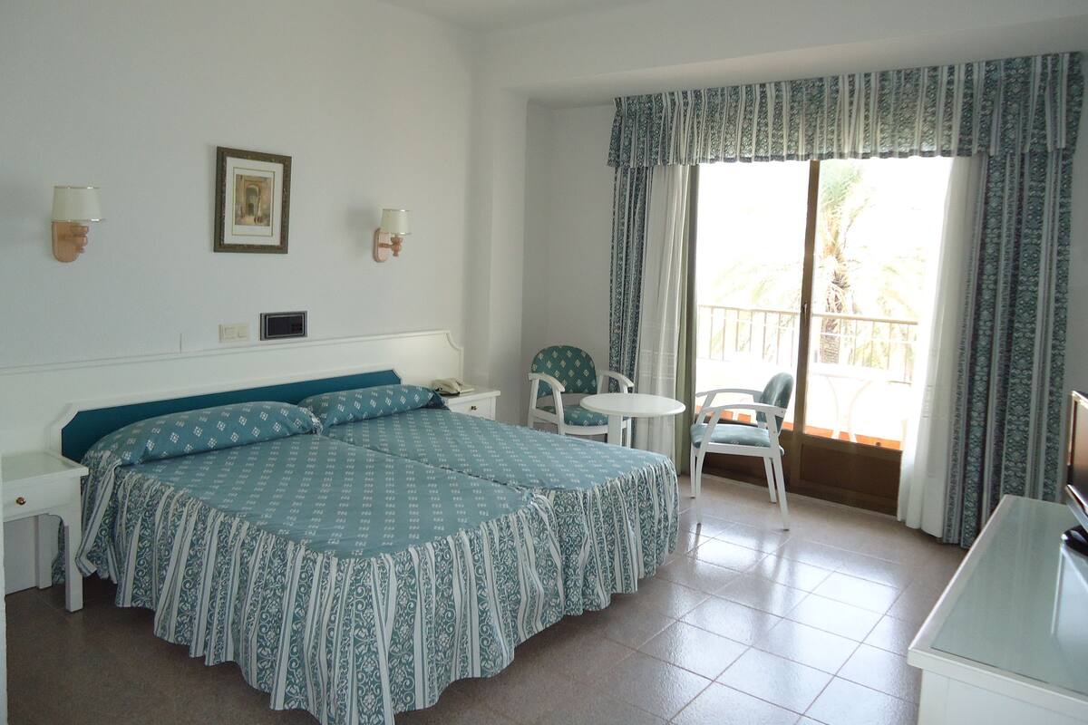 Une chambre d’hôtel avec un grand lit, une petite table, deux chaises, un bureau et un balcon avec des rideaux.
