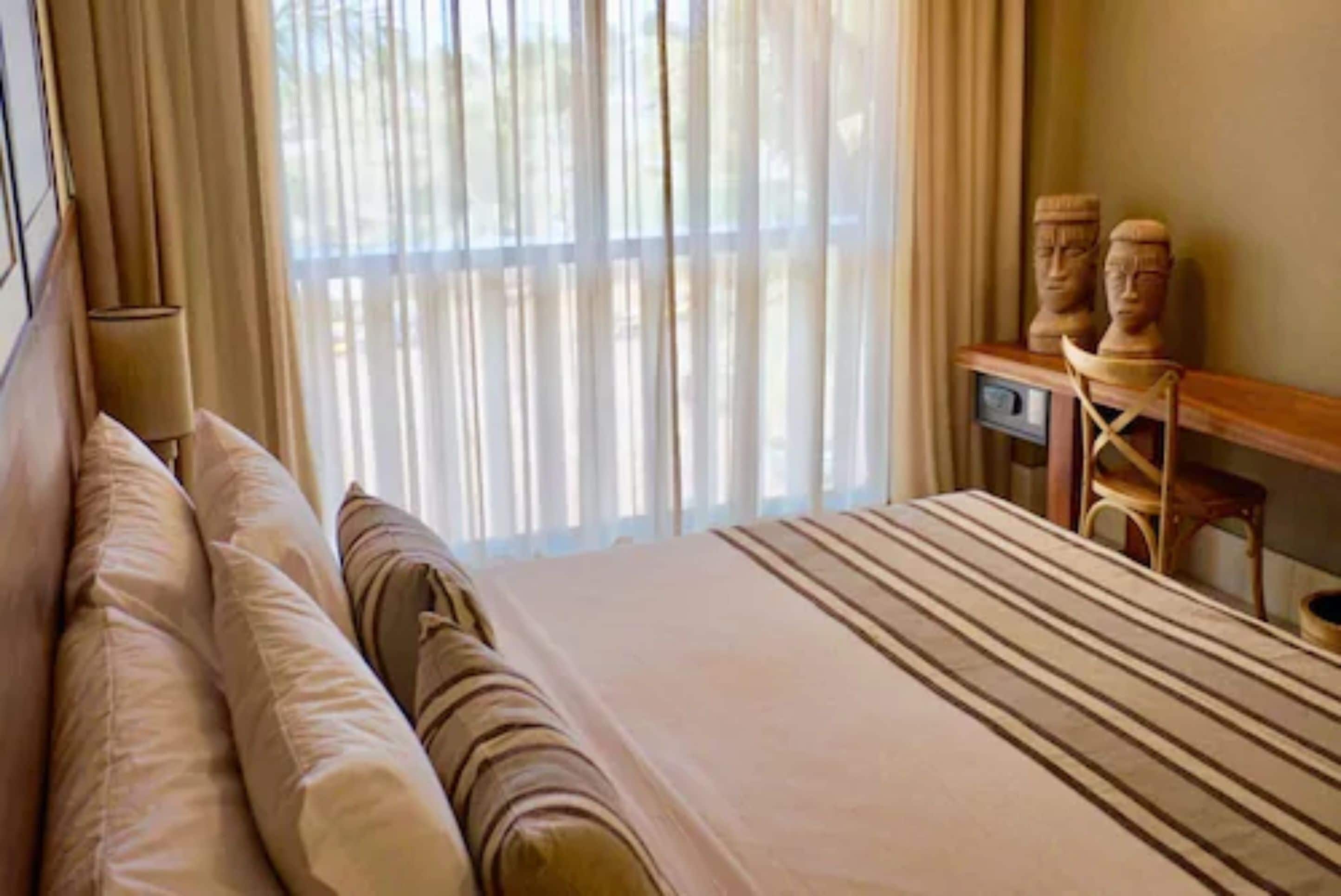 A hotel room with a large bed, a wooden nightstand, and a window with sheer curtains.