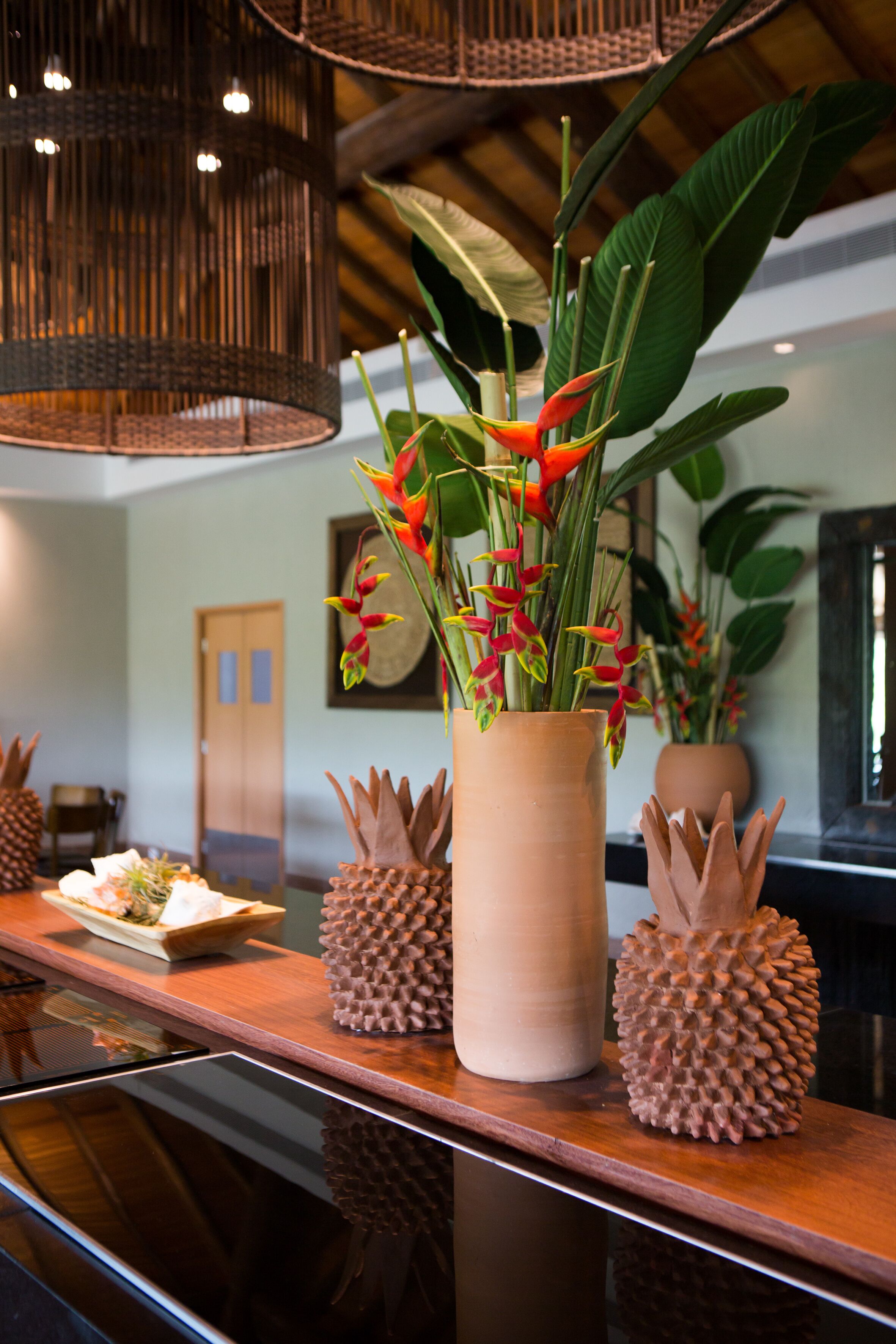 A reception area with a wooden counter, a large vase with tropical flowers, and a woven light fixture.