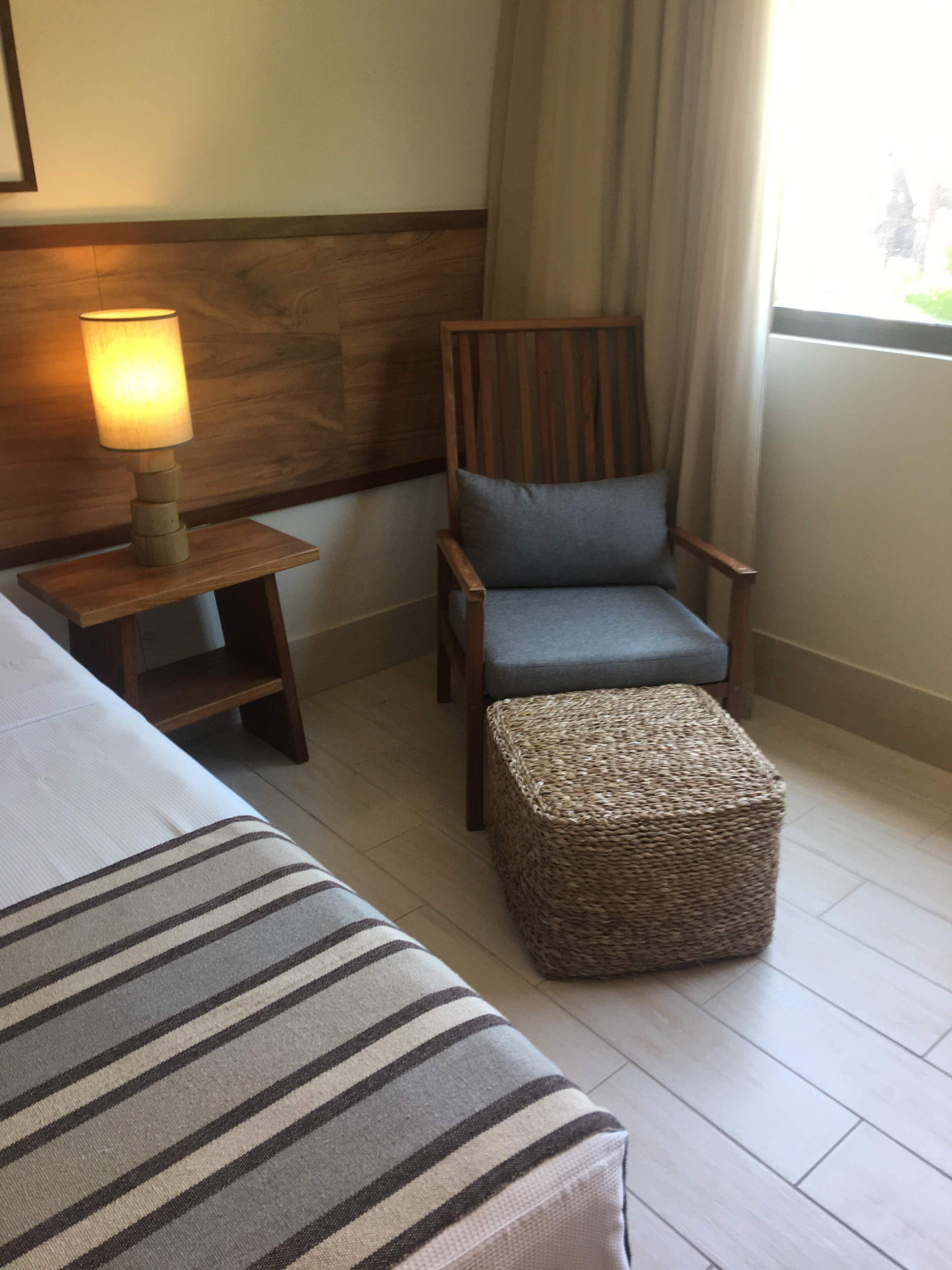 A hotel room with a bed, a chair with a cushion, a small table with a lamp, and a woven ottoman.