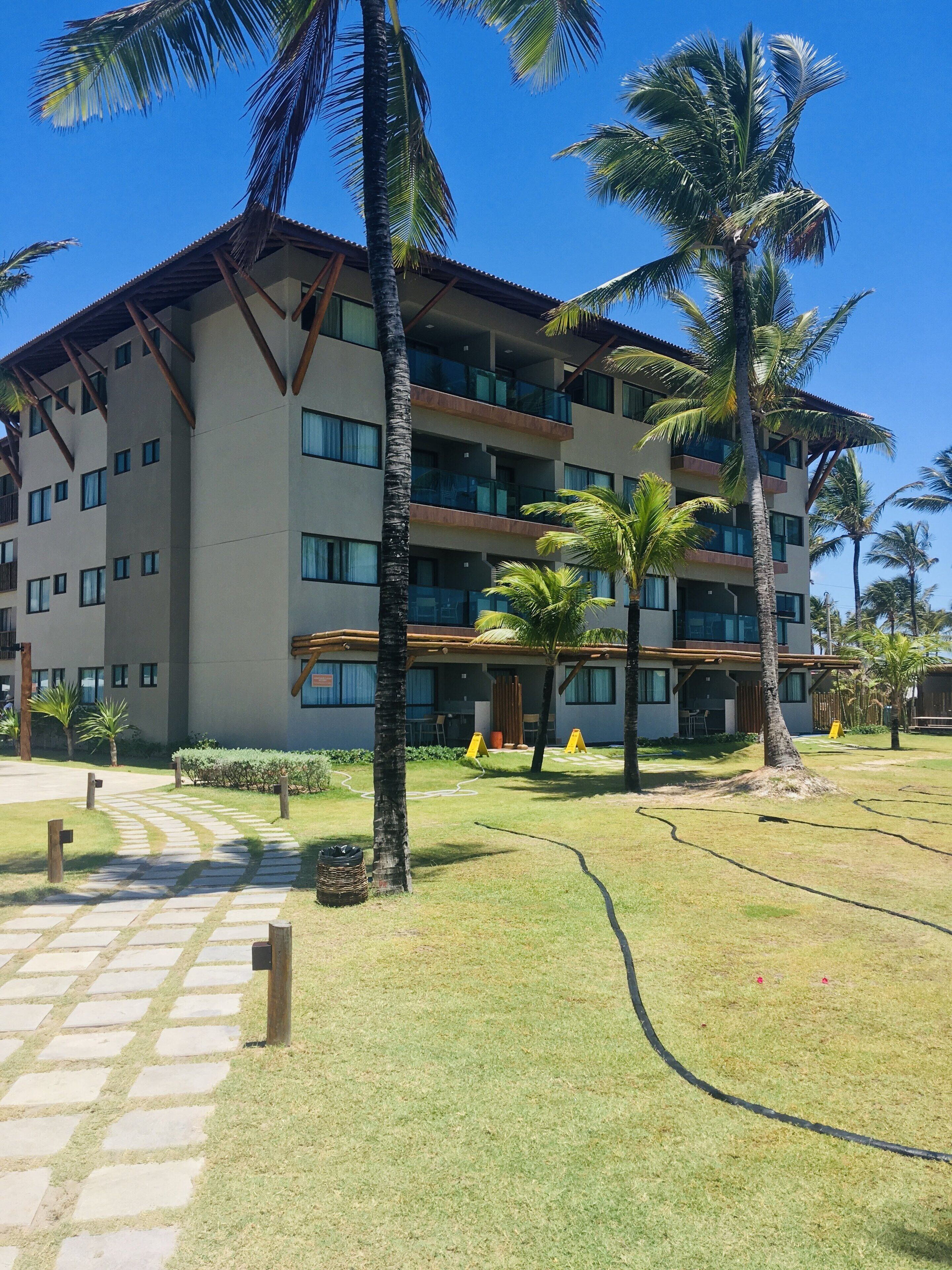 Un hotel de varios pisos con techo plano, amplias ventanas y un patio pavimentado con palmeras.
