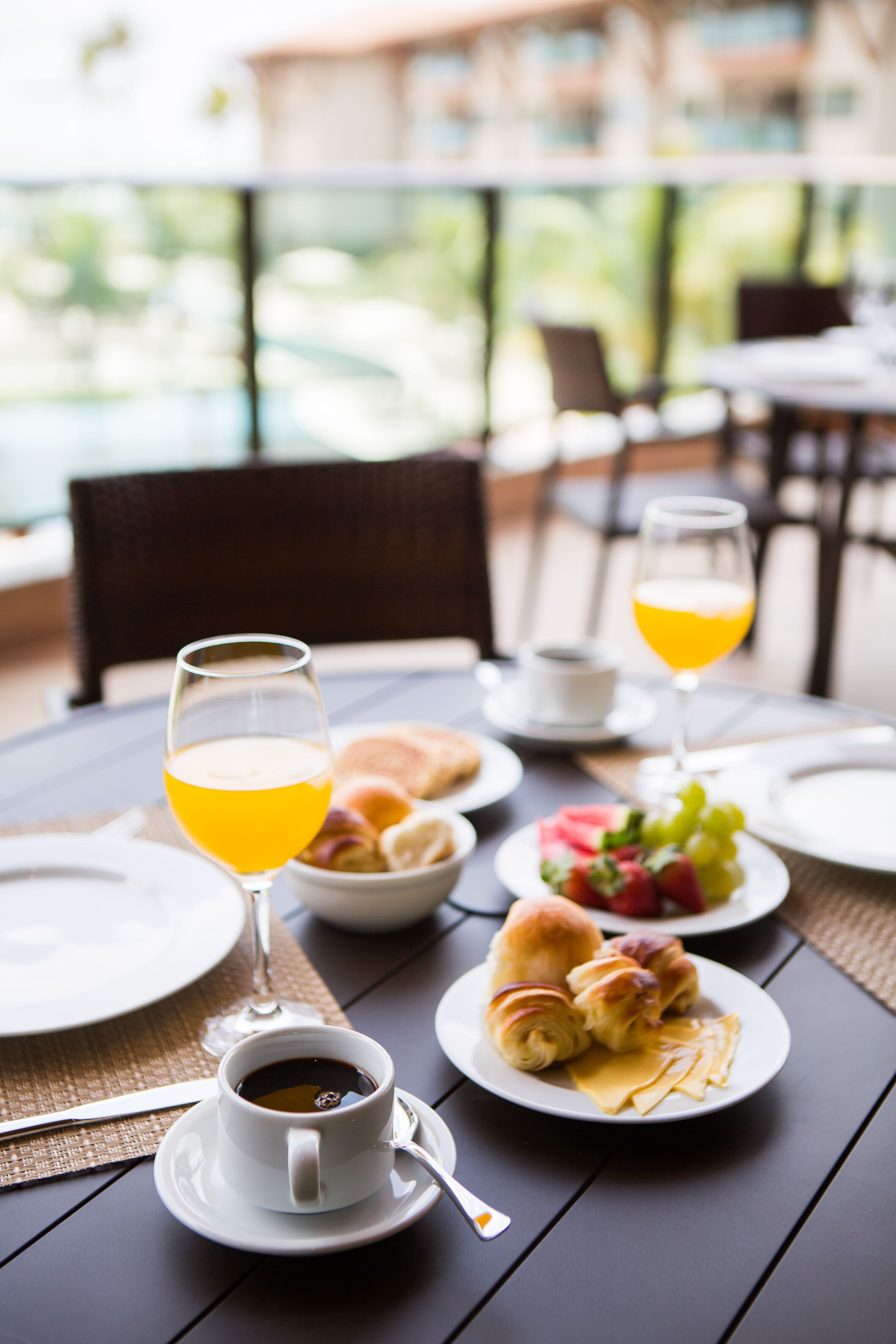 Et spisebord med morgenmadsprodukter, herunder croissanter, frugt og drikkevarer, placeret på en udendørs terrasse med udsigt til en pool og grøn bevoksning.