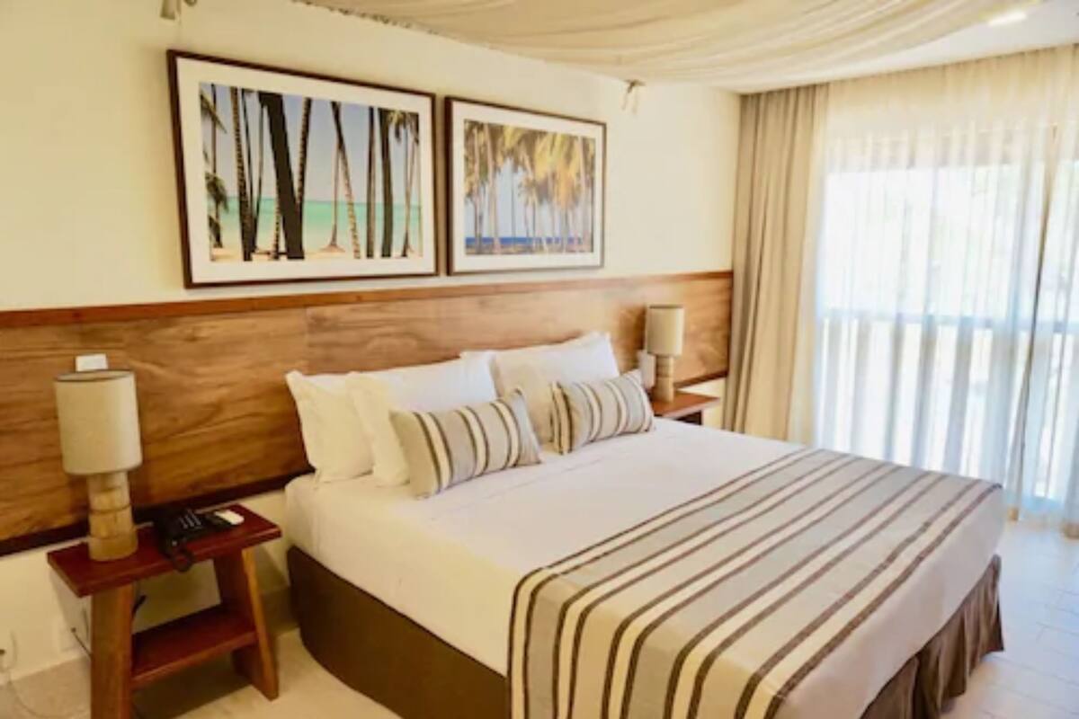 A hotel room with a large bed, wooden headboard, two framed pictures on the wall, a nightstand with a lamp, and a window with curtains.