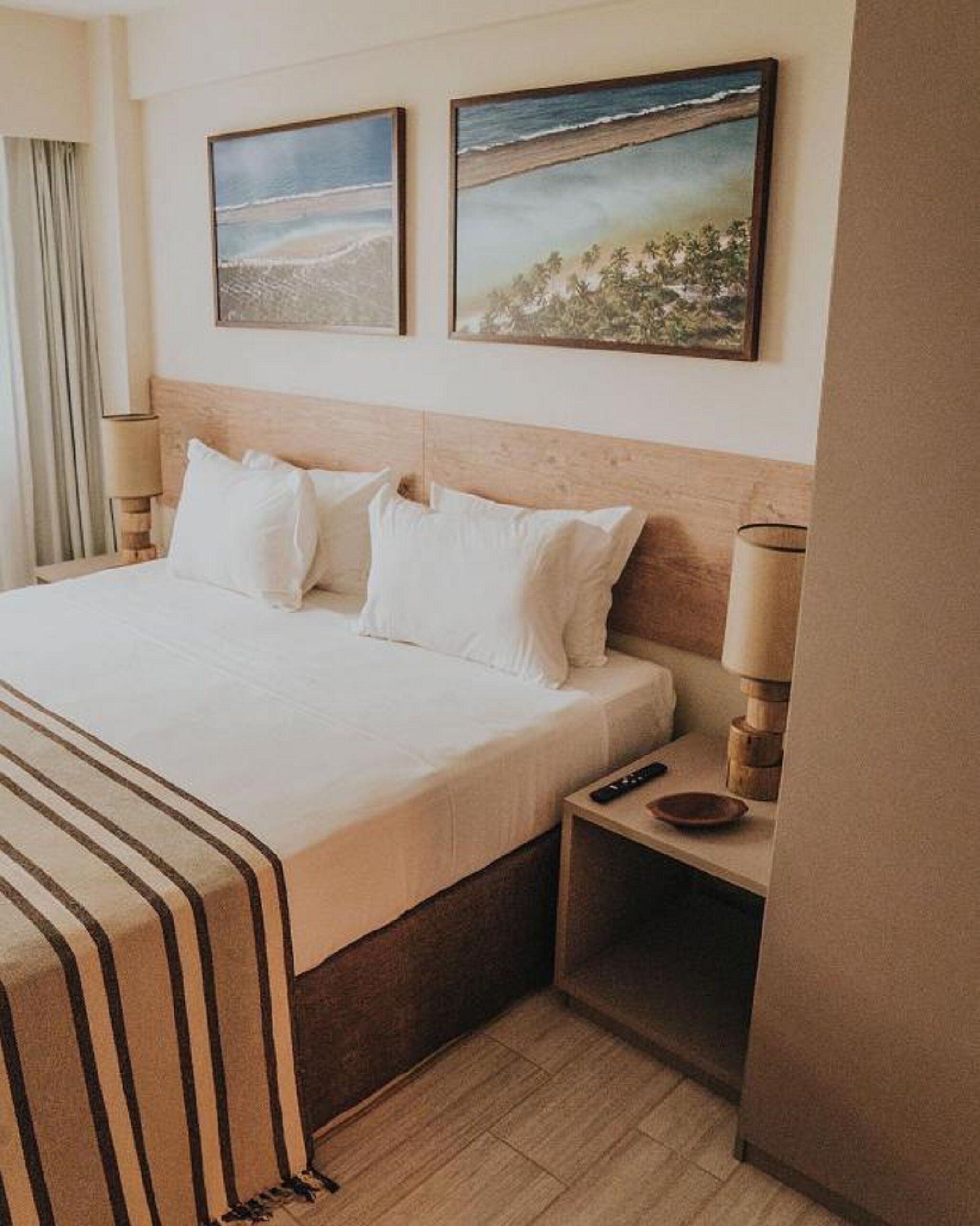 A hotel room with a large bed, two wall-mounted paintings, a bedside table with a lamp, and a remote control.