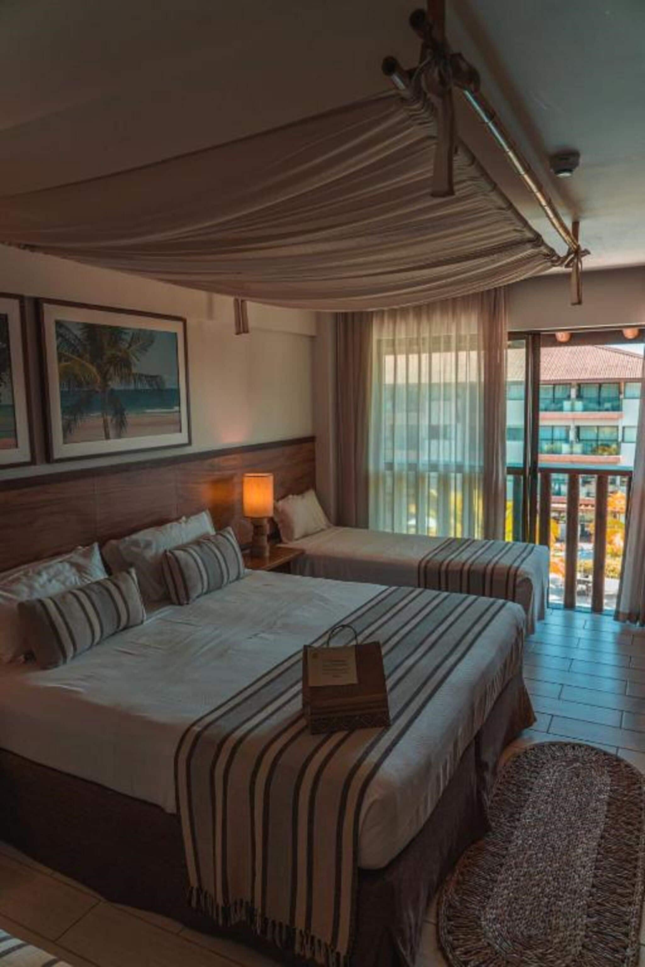 A hotel room with a large bed, a smaller bed, a nightstand with a lamp, a ceiling fan, and a view of the outside.