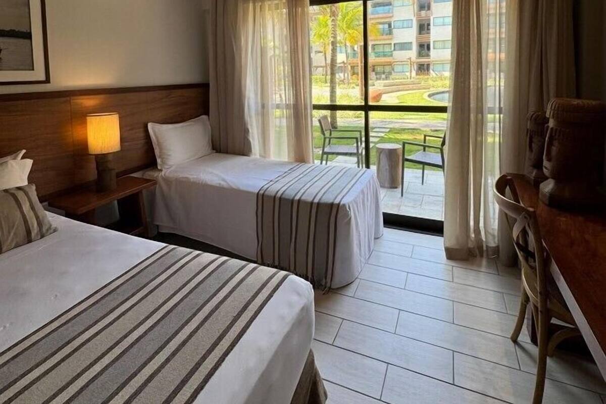 A hotel room with two beds, a nightstand with a lamp, a chair, and a view of a building and greenery outside.