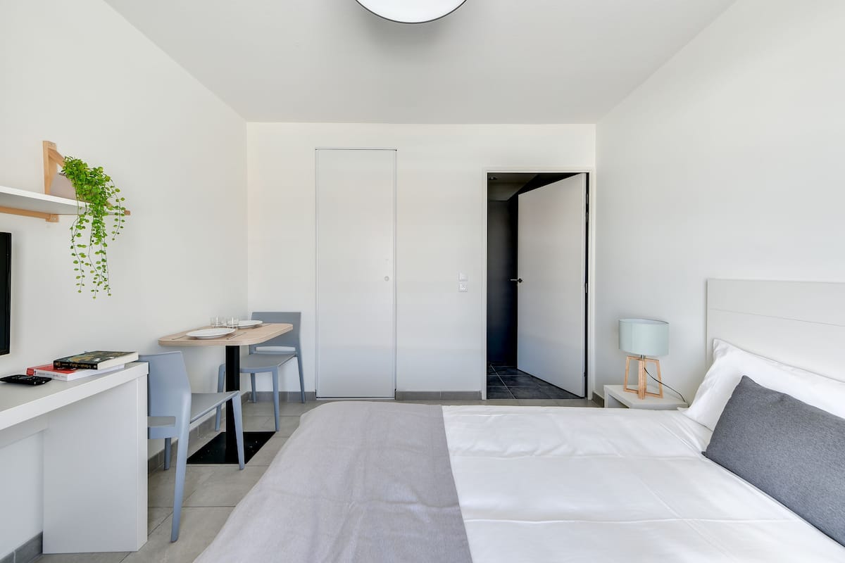 Une chambre d’hôtel moderne, équipée d’un lit, d’un bureau, d’une chaise, d’une petite table, d’une lampe et d’une plante.