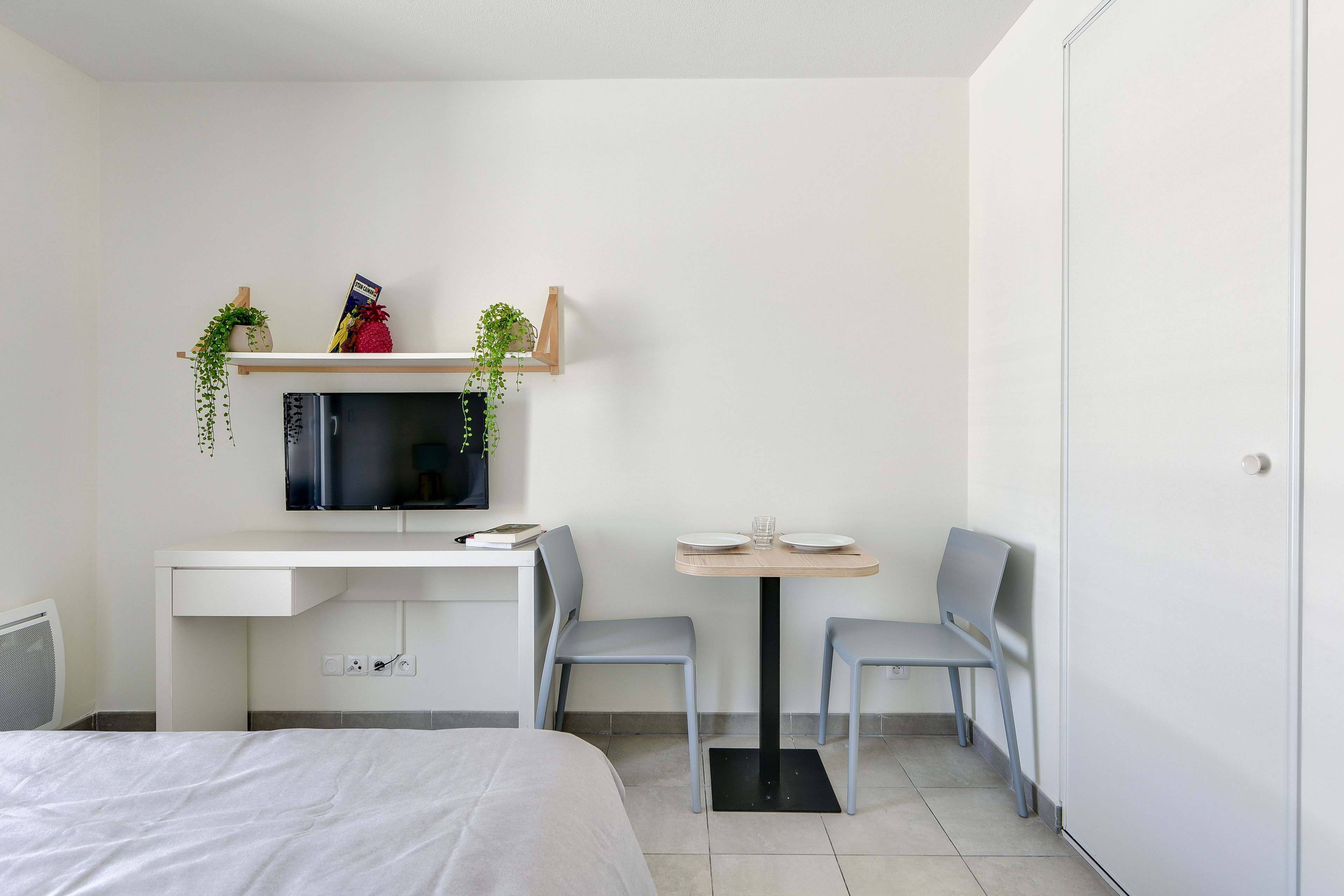 A hotel room with a bed, a desk with a TV, a small table with chairs, and a door.