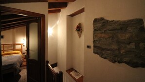 A narrow hallway with a wooden door leading to a bedroom with a bed and a nightstand. A stone wall with a decorative flower arrangement is visible on the right. - b&b Vecchio Torchio (Bard)