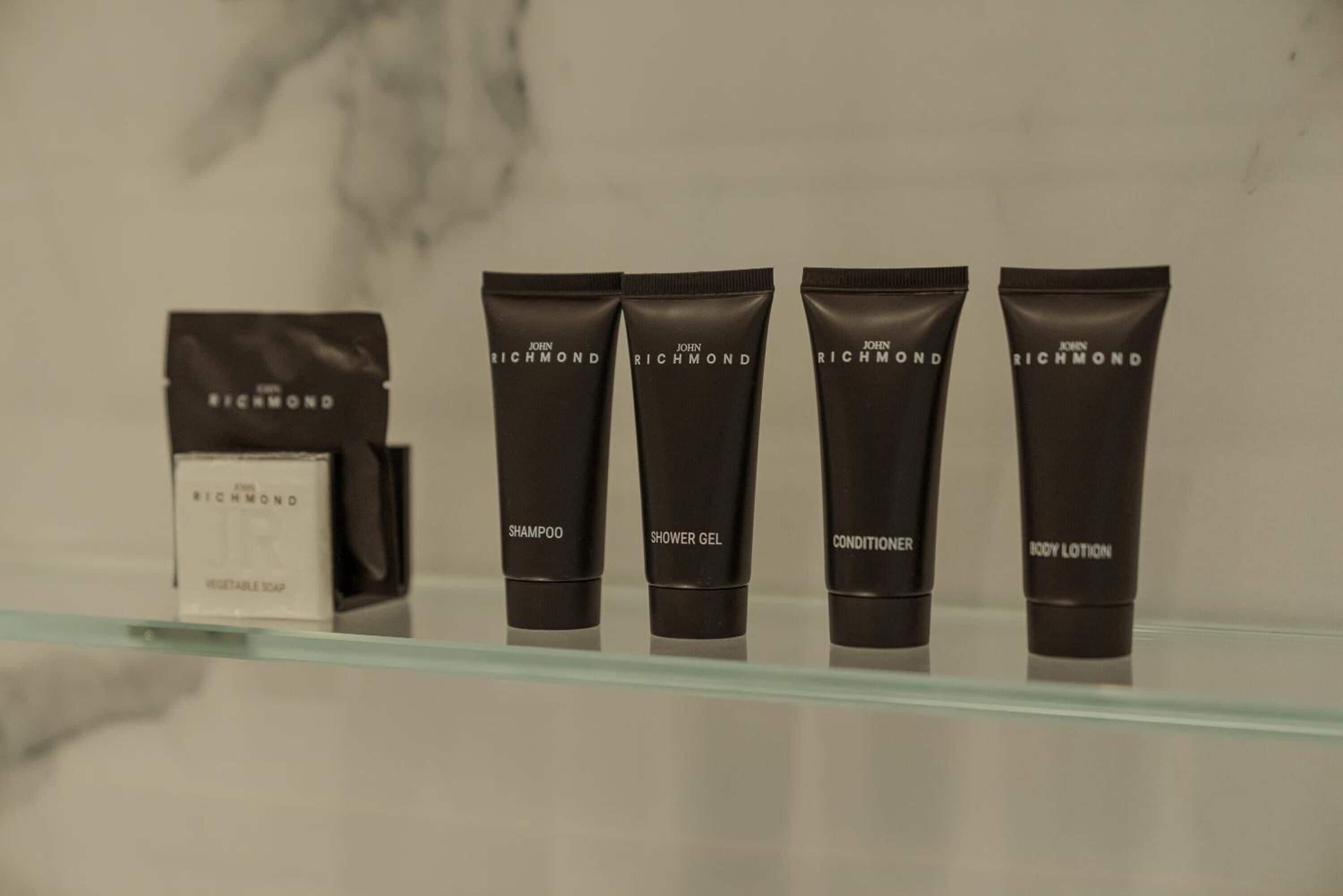 Exhibición de productos John Richmond que incluye champú, gel de ducha, acondicionador y loción corporal sobre una repisa de vidrio.