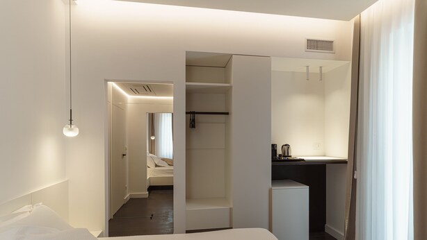 Ein modernes Hotelzimmer mit einem großen Bett, einem begehbaren Kleiderschrank, einer kleinen Küchenzeile und einem durchgehenden Türrahmen einsehbarer Badezimmer.