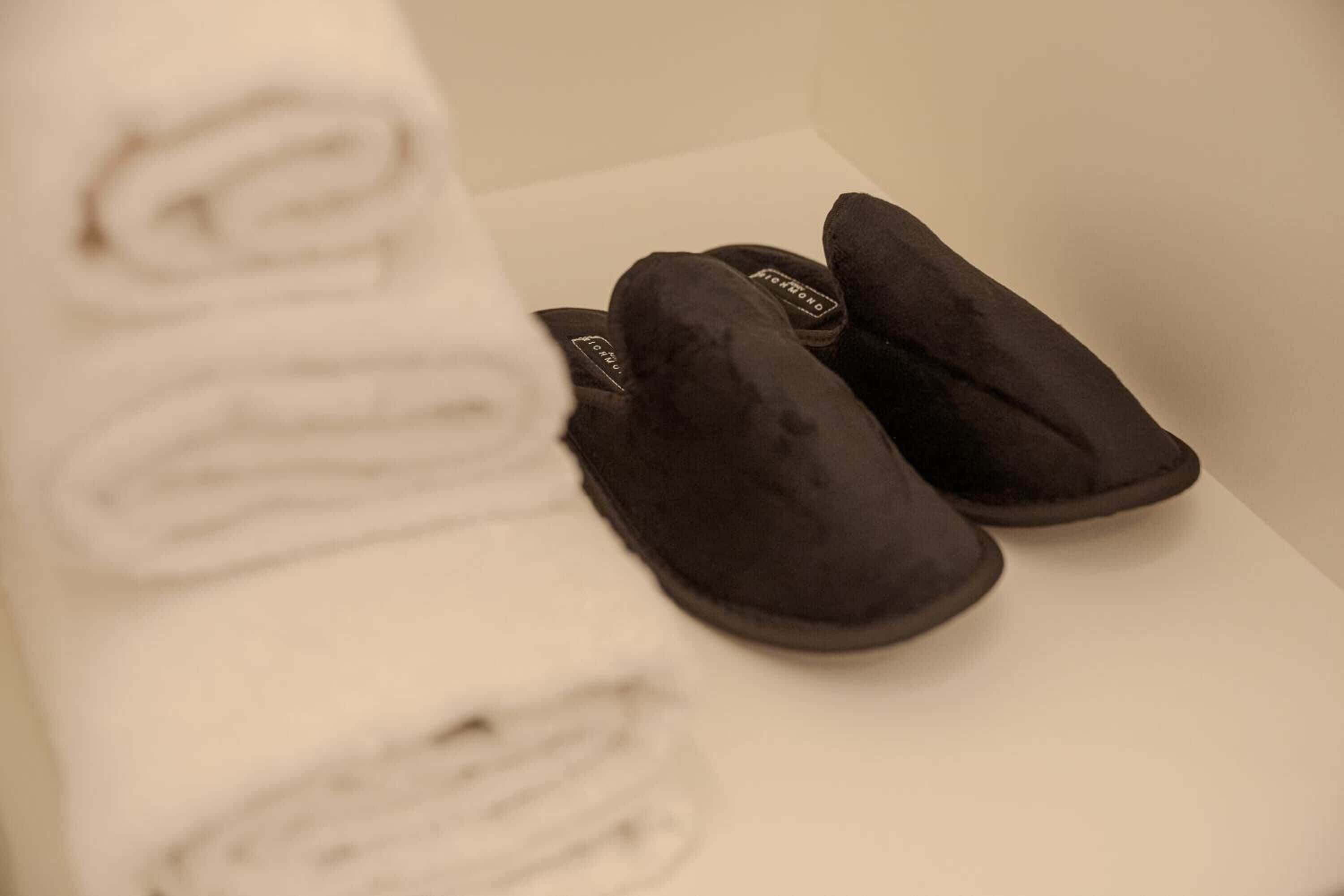 Zwarte pantoffels en een opgerolde witte handdoek op een witte ondergrond.