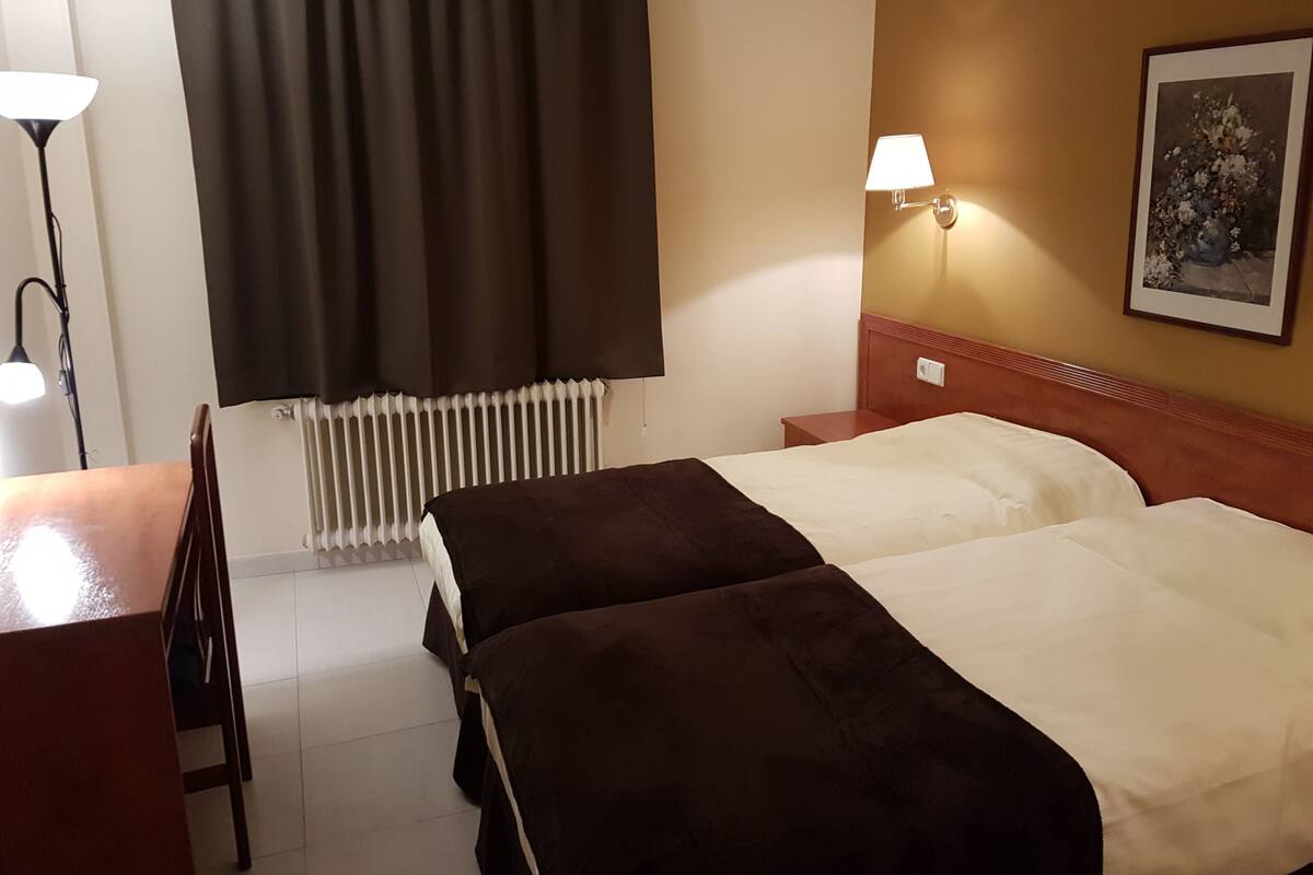 A hotel room with two beds, a desk, a chair, a radiator, and a wall-mounted lamp.
