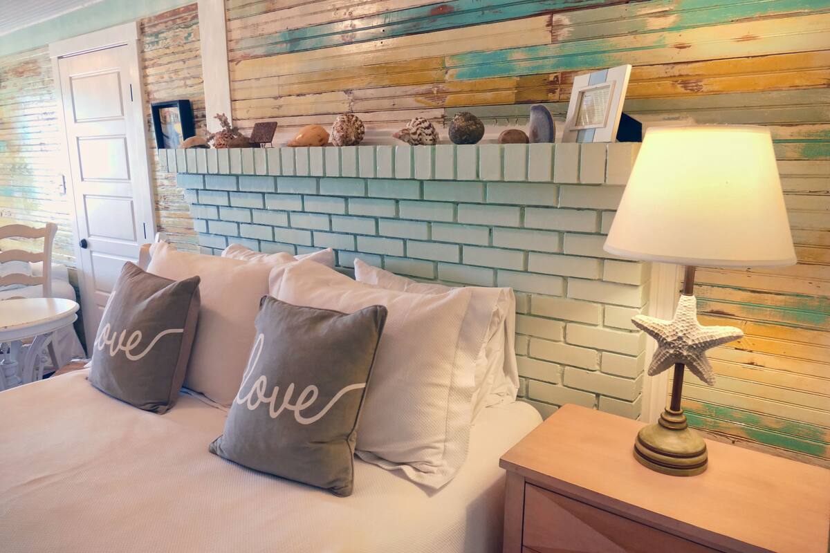 A bedroom with a brick wall, a bed with pillows, a nightstand, and a lamp.