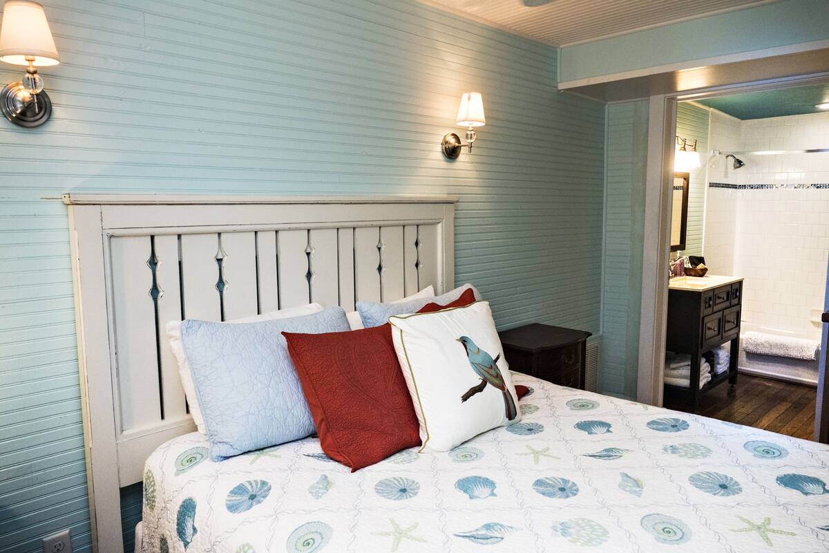 A bedroom with a bed, pillows, and a nightstand. There is a bathroom visible in the background.
