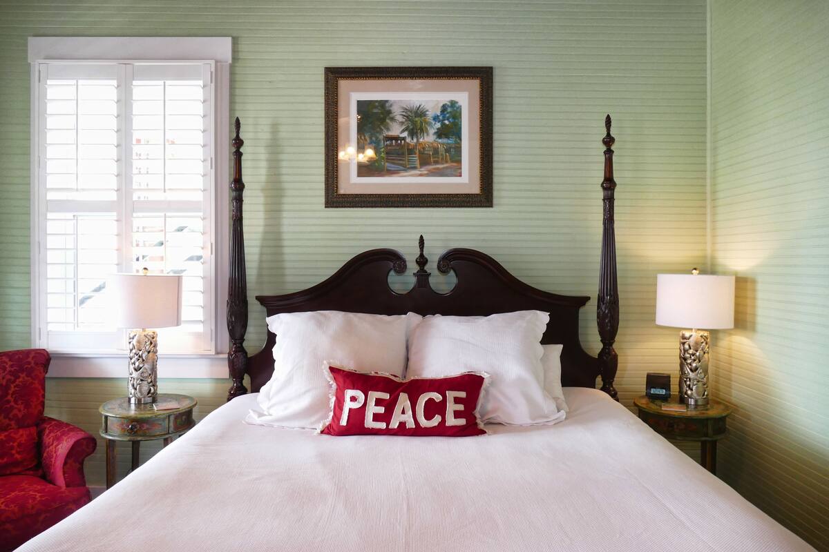 A neatly made bed with a pillow labeled "PEACE," a framed picture on the wall, and a window with blinds.