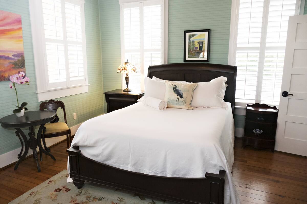 A bedroom with a large bed, a nightstand with a lamp, a chair, and a small table.