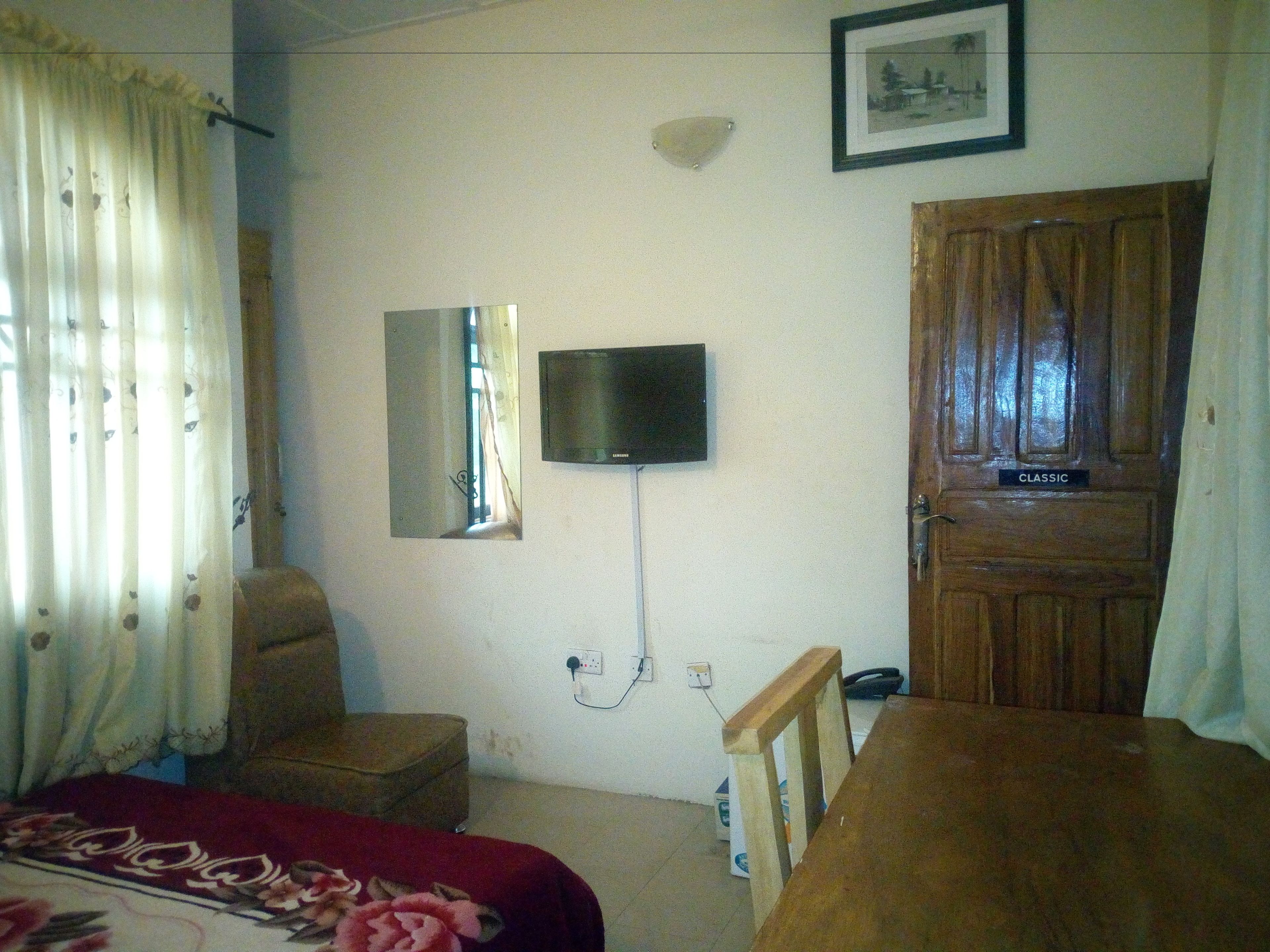 A room with a bed, a TV mounted on the wall, a wooden door labeled "CLASSIC", and a window with curtains.