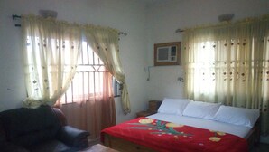 A bedroom with a bed, a wooden nightstand, a chair, and curtains. - Blue Banana Resorts (Abeokuta)