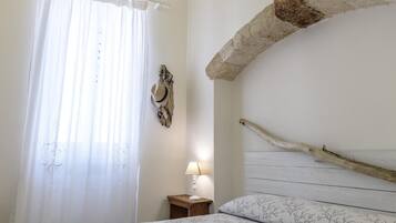 A bedroom with a bed, a nightstand, a lamp, a window with curtains, and a rustic stone beam.