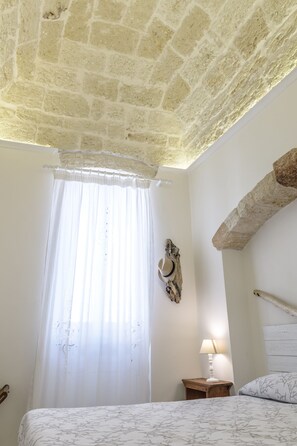A bedroom with a stone ceiling, a bed with a patterned bedspread, a nightstand with a lamp, and a rustic wall decor piece. - Casa Meliò (Polignano a Mare)