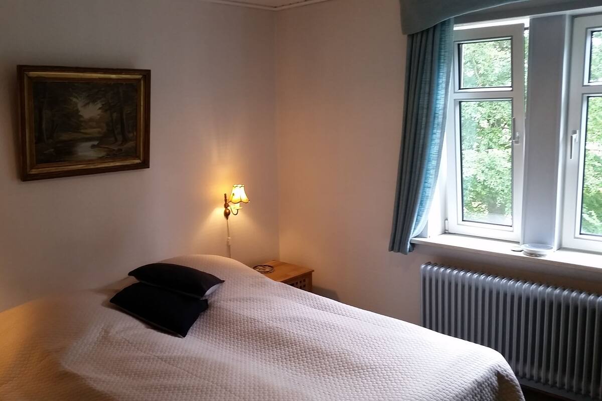 A bedroom with a bed, a nightstand, a lamp, a painting, and a window with curtains.