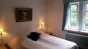 Une chambre à coucher avec un lit, une table de chevet, une lampe, un tableau et une fenêtre avec des rideaux.