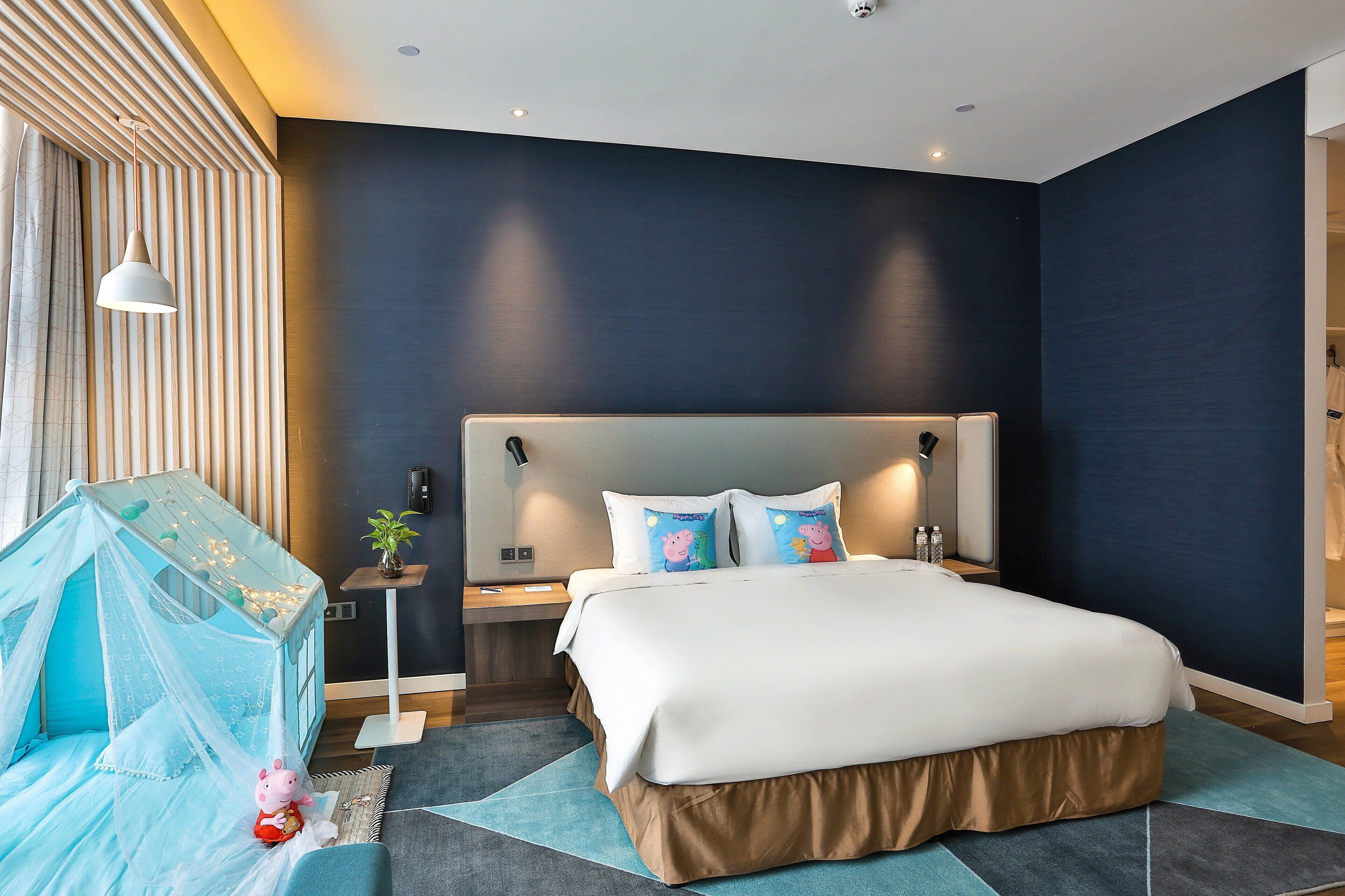 A modern hotel room with a large bed, bedside tables, a nightstand, a bedside lamp, a wall-mounted phone, and a window with blinds.