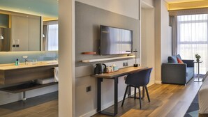 A modern hotel room with a flat-screen TV mounted on the wall, a wooden desk with a chair, a sofa, and a small side table with a plant. - Holiday Inn Express Harbin Qunli by IHG (Harbin)