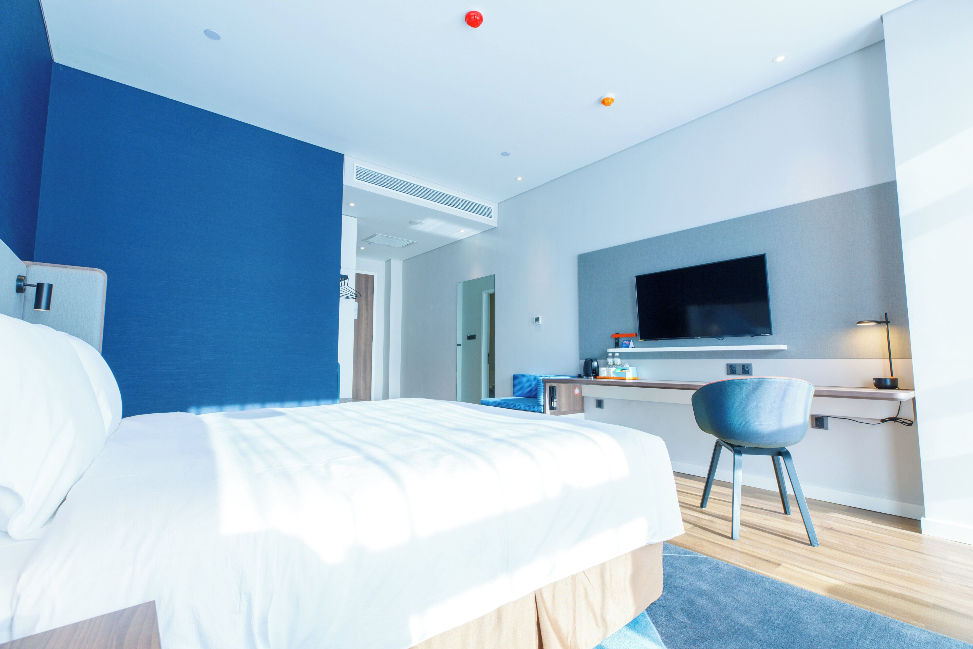 A modern hotel room with a large bed, a desk, a chair, a flat-screen TV, and a bathroom visible in the background.