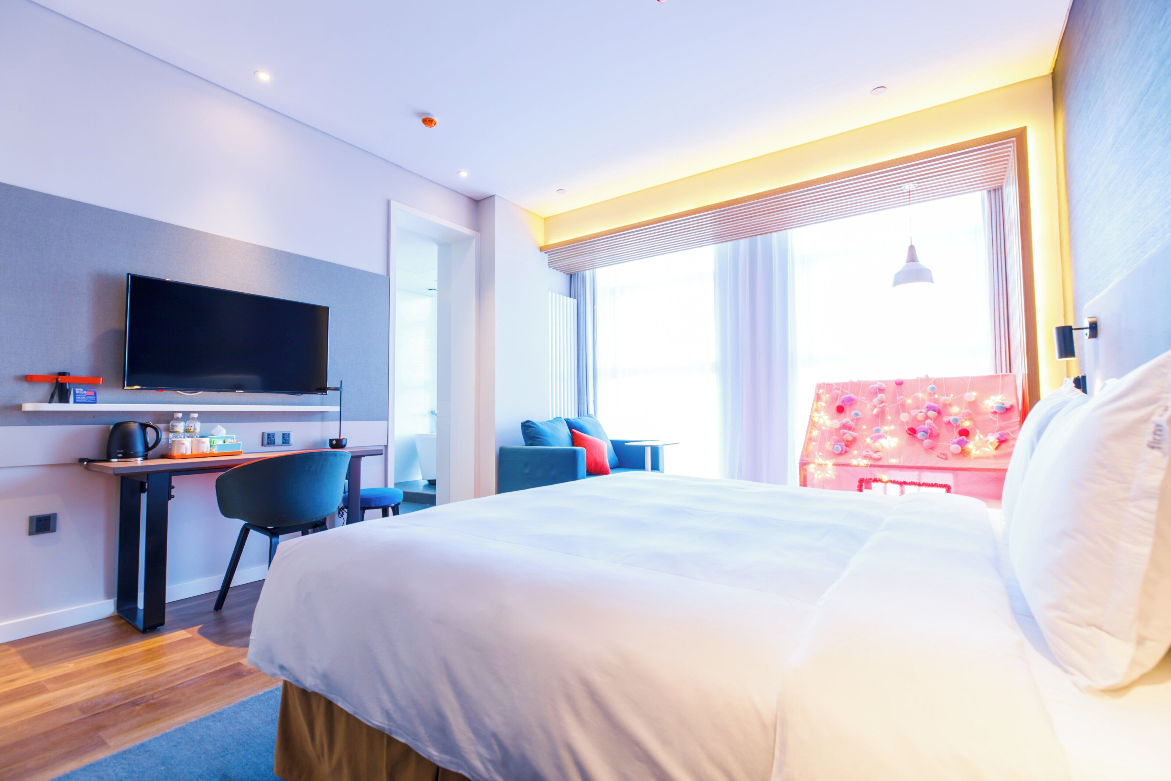 Habitación de hotel moderna con una cama grande, un escritorio con televisor de pantalla plana, una zona de estar con sillones y una ventana con cortinas.