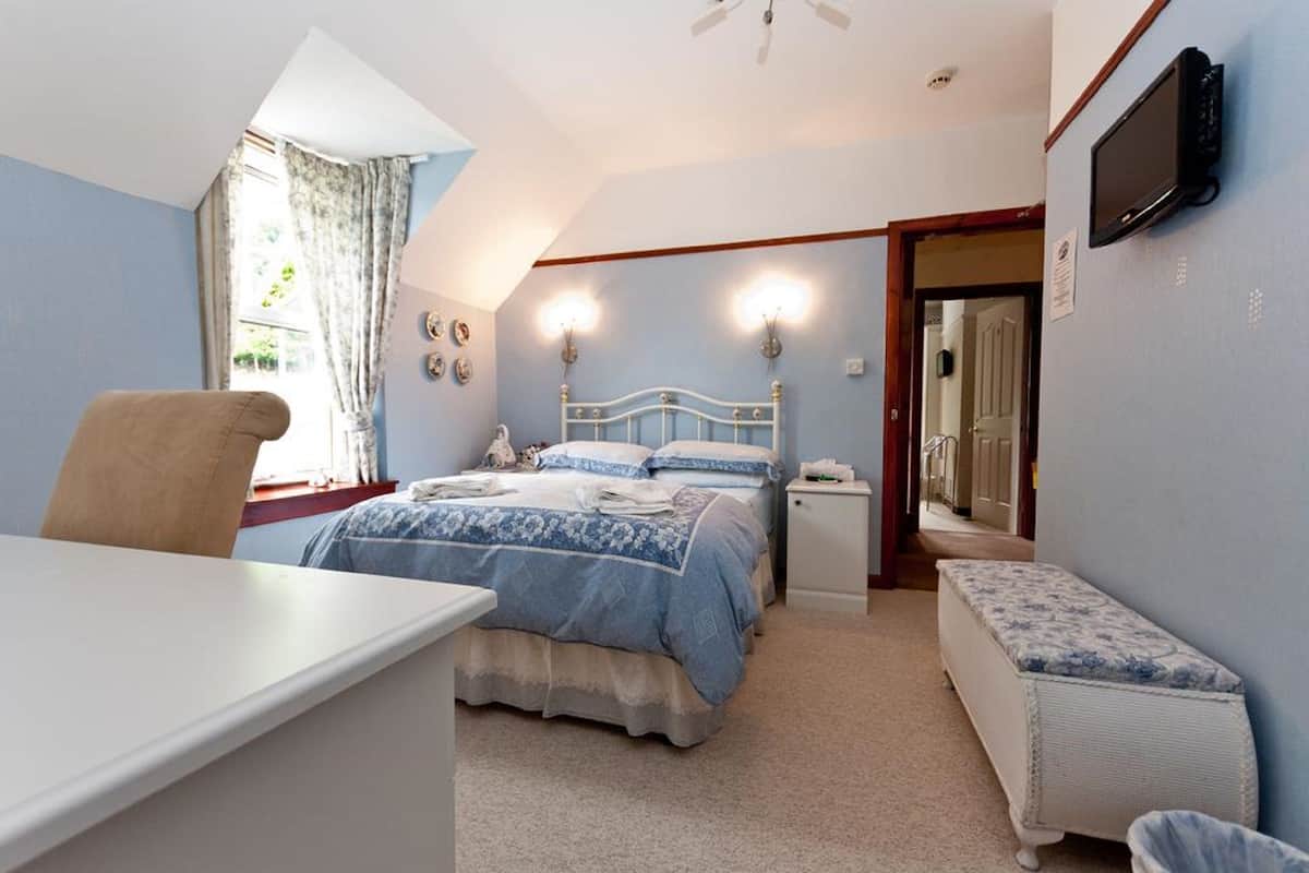 A bedroom with a bed, bedside tables, a desk, a chair, a bench, a ceiling fan, and a TV mounted on the wall.