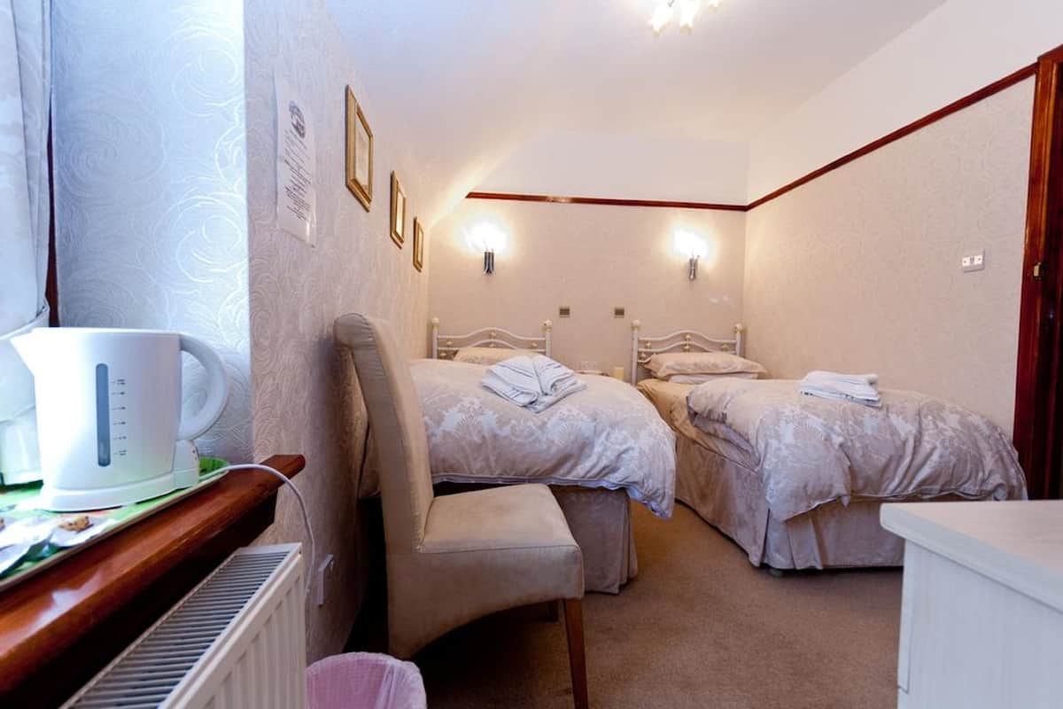 A hotel room with two beds, a desk with a kettle, and a chair.