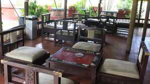 Een terras met houten meubels in klassieke stijl, inclusief zachte stoelen en een centrale tafel, omgeven door potplanten en een betegelde vloer.