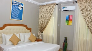 A neatly made bed with white and beige bedding, a poster on the wall, and a vase on the floor. - Jacaranda Suites (Calabar)