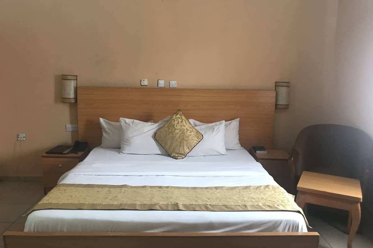 Ein ordentlich bezogenes Bett mit weißen Leinentextilien und einem dekorativen Kissen, ein Nachttisch mit Lampe sowie ein Stuhl in einem schlichten Zimmer mit beige gestrichenen Wänden.