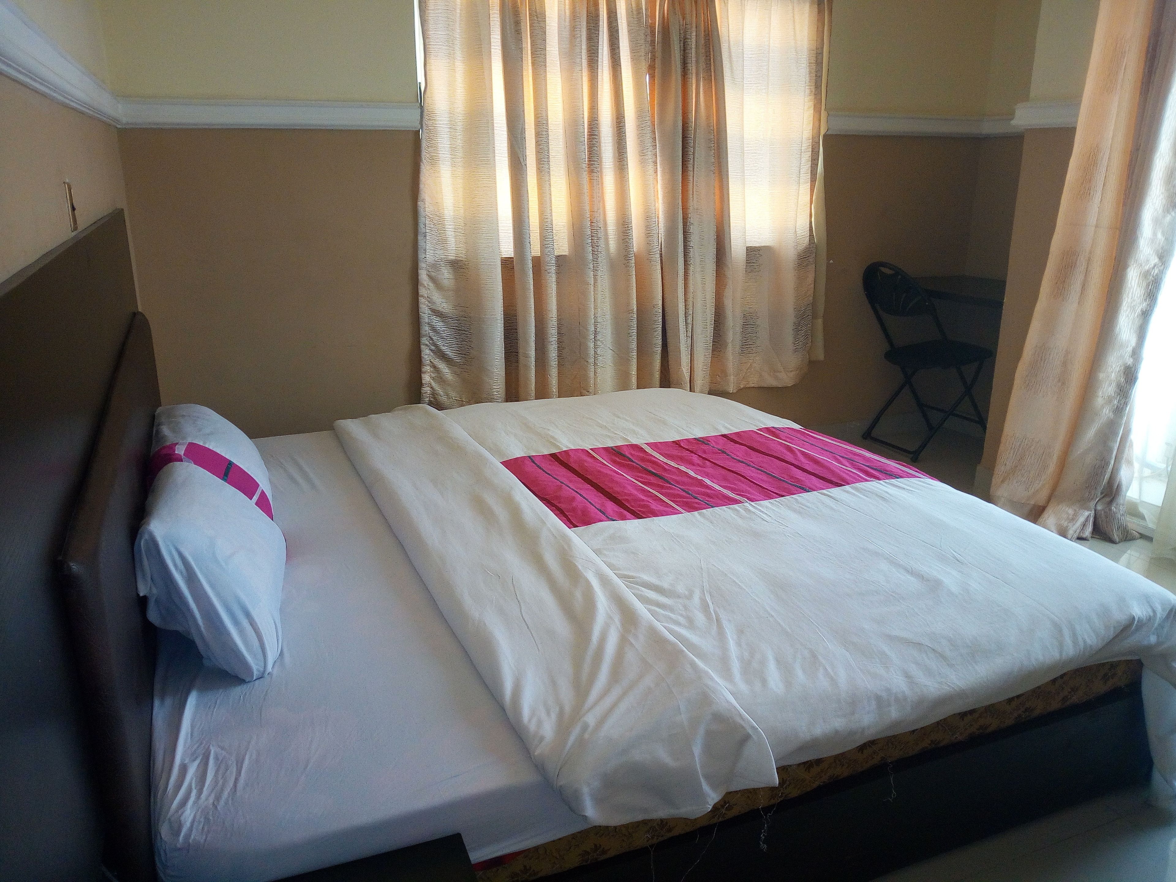 Una cama bien hecha con sábanas blancas y un detalle en rosa, una silla y una ventana con cortinas de encaje.