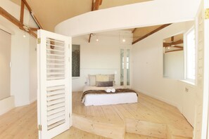 Une chambre avec un plancher en bois, un lit recouvert d’une courtepointe blanche et une grande fenêtre.