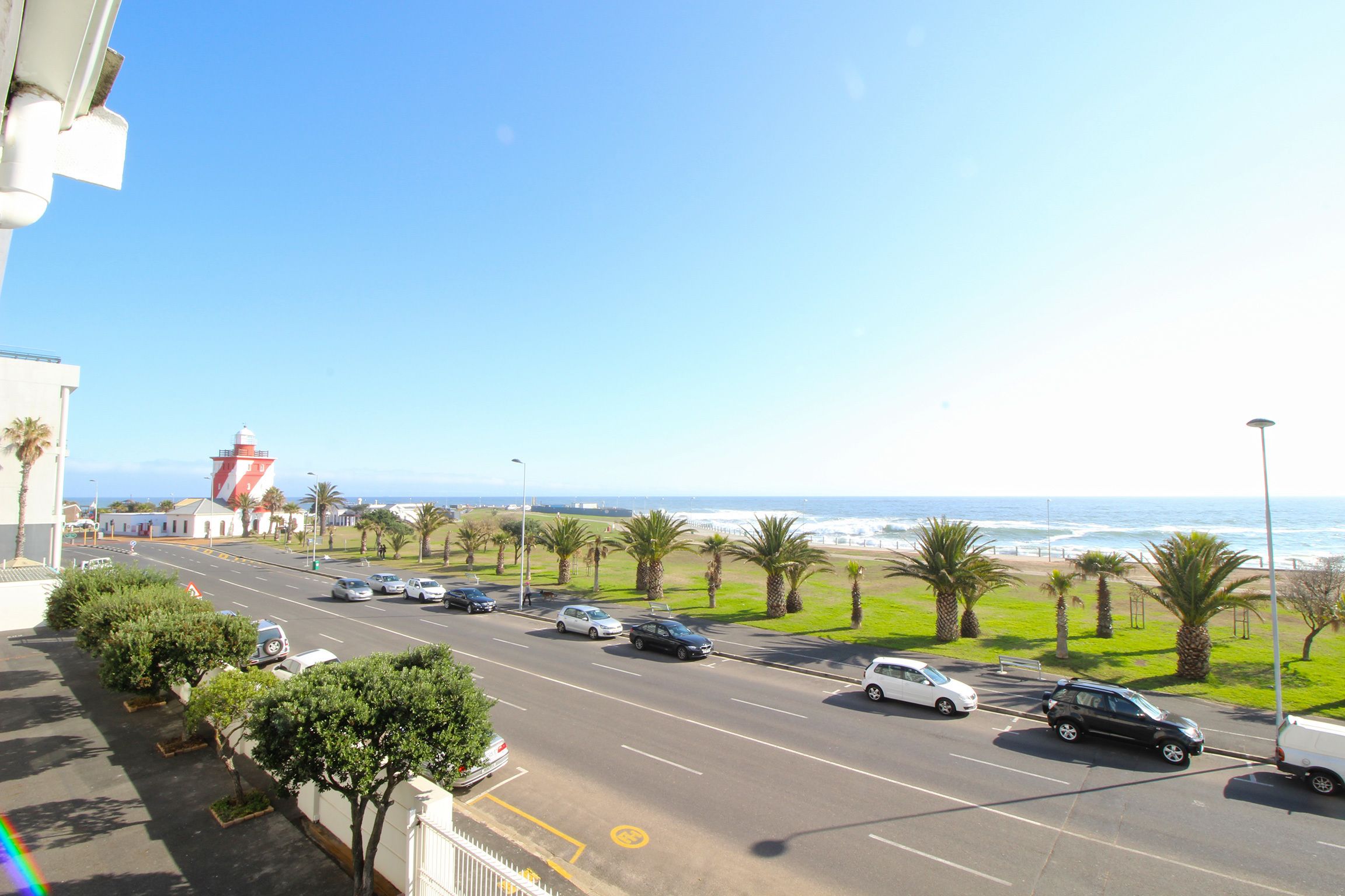 A coastal road with parked cars, a lighthouse, and a view of the ocean.