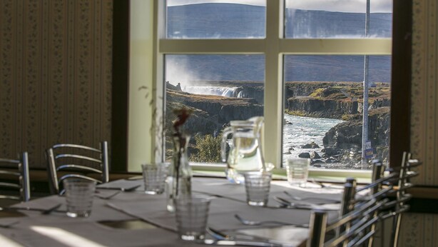A dining table with chairs, a pitcher, and glasses, set against a window with a view of a waterfall and mountains. - Fosshóll Guesthouse (Laugar)