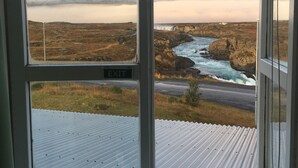 A view from a window with an 'EXIT' sign, overlooking a coastal landscape with a river, cliffs, and a road. - Fosshóll Guesthouse (Laugar)