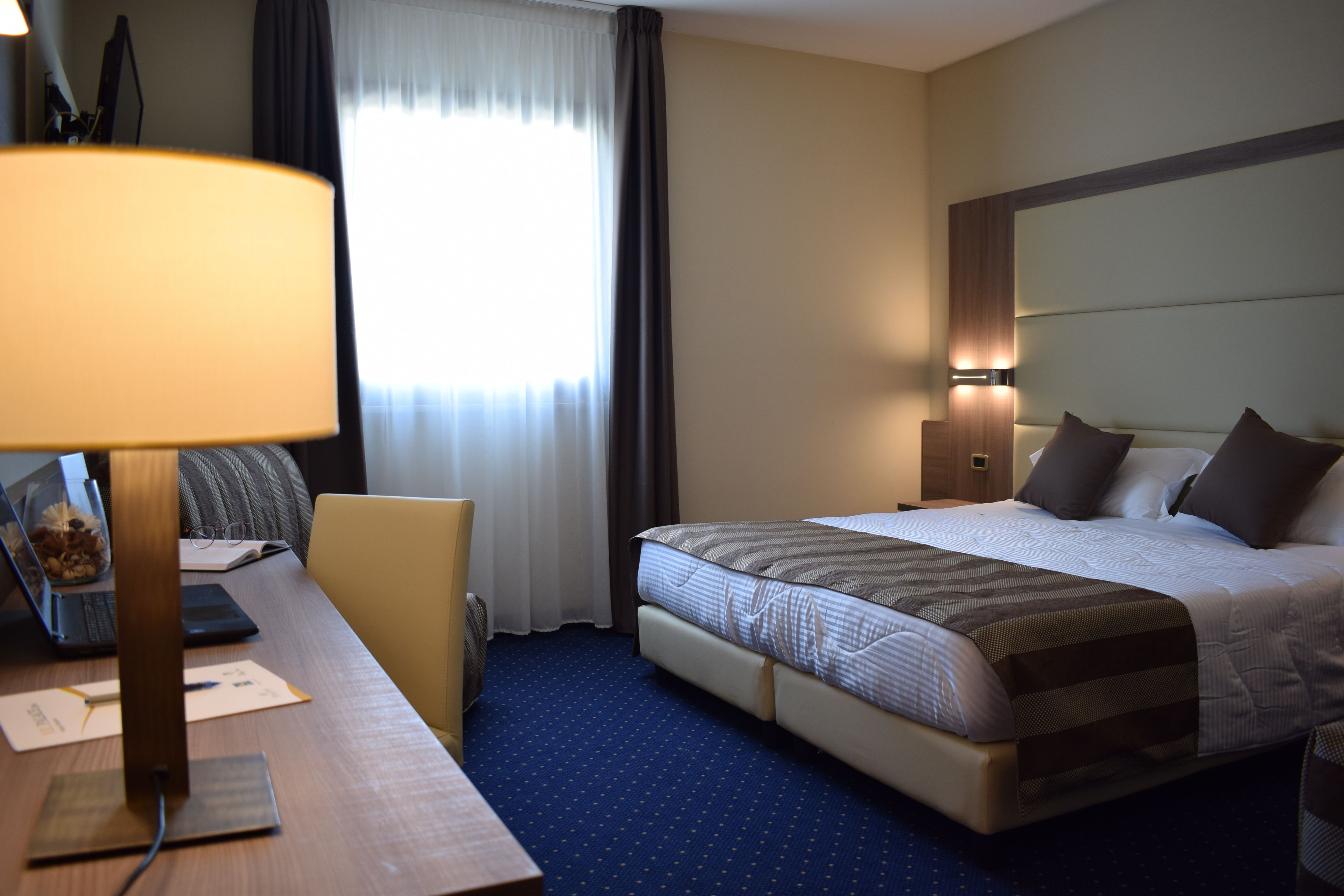 Habitación de hotel con una cama, un escritorio con lámpara, una silla y una ventana con cortinas.