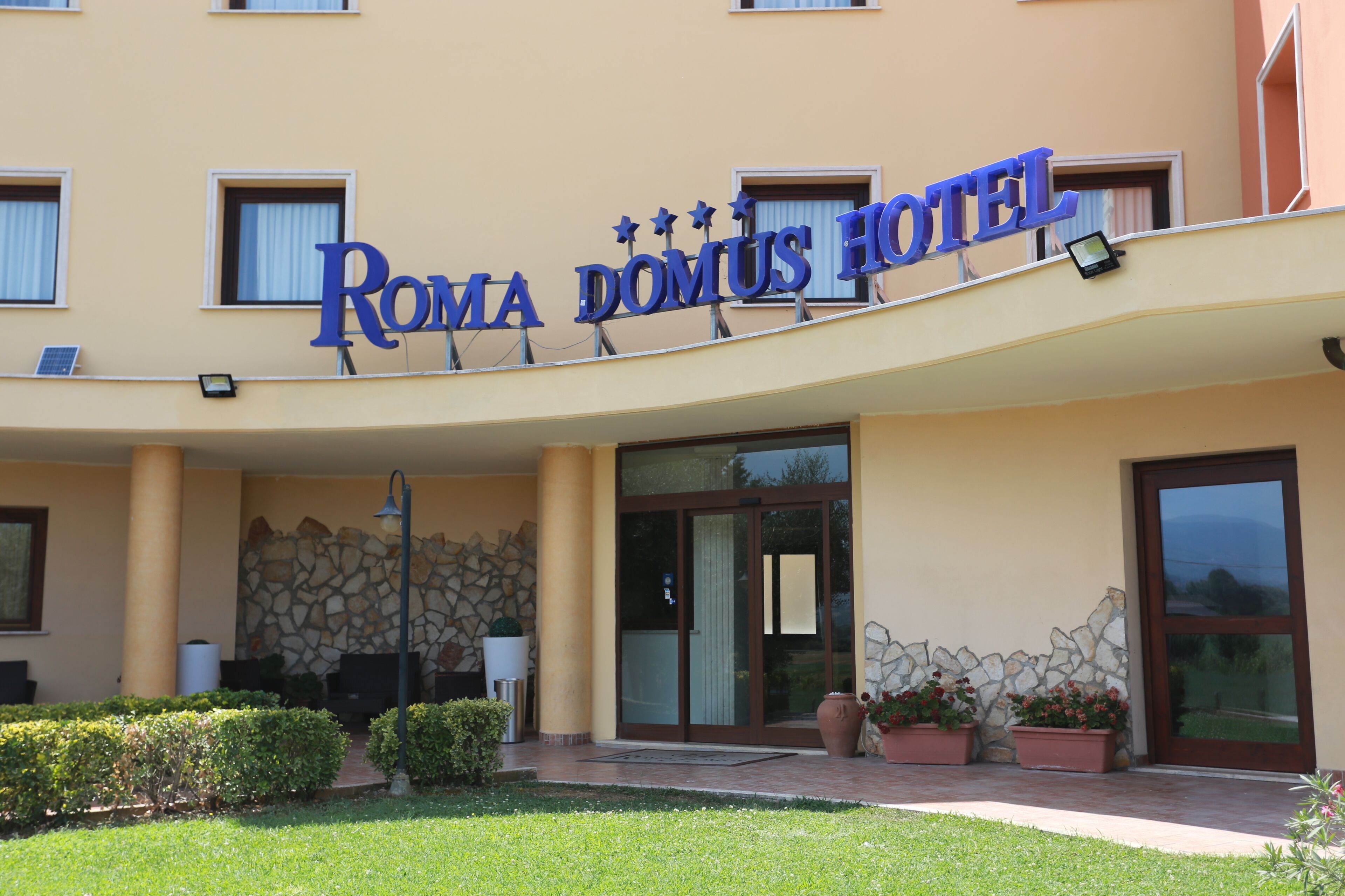The exterior of the Roma Domus Hotel with a stone facade, large glass entrance doors, and potted plants.